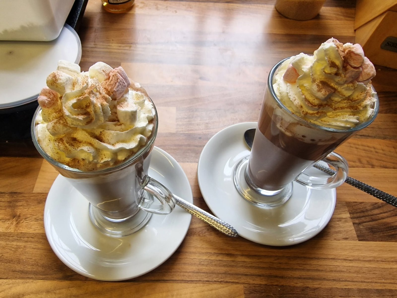 Two hot drinks in tall glass mugs on saucers, topped with whipped cream, marshmallows and a dusting of cinnamon